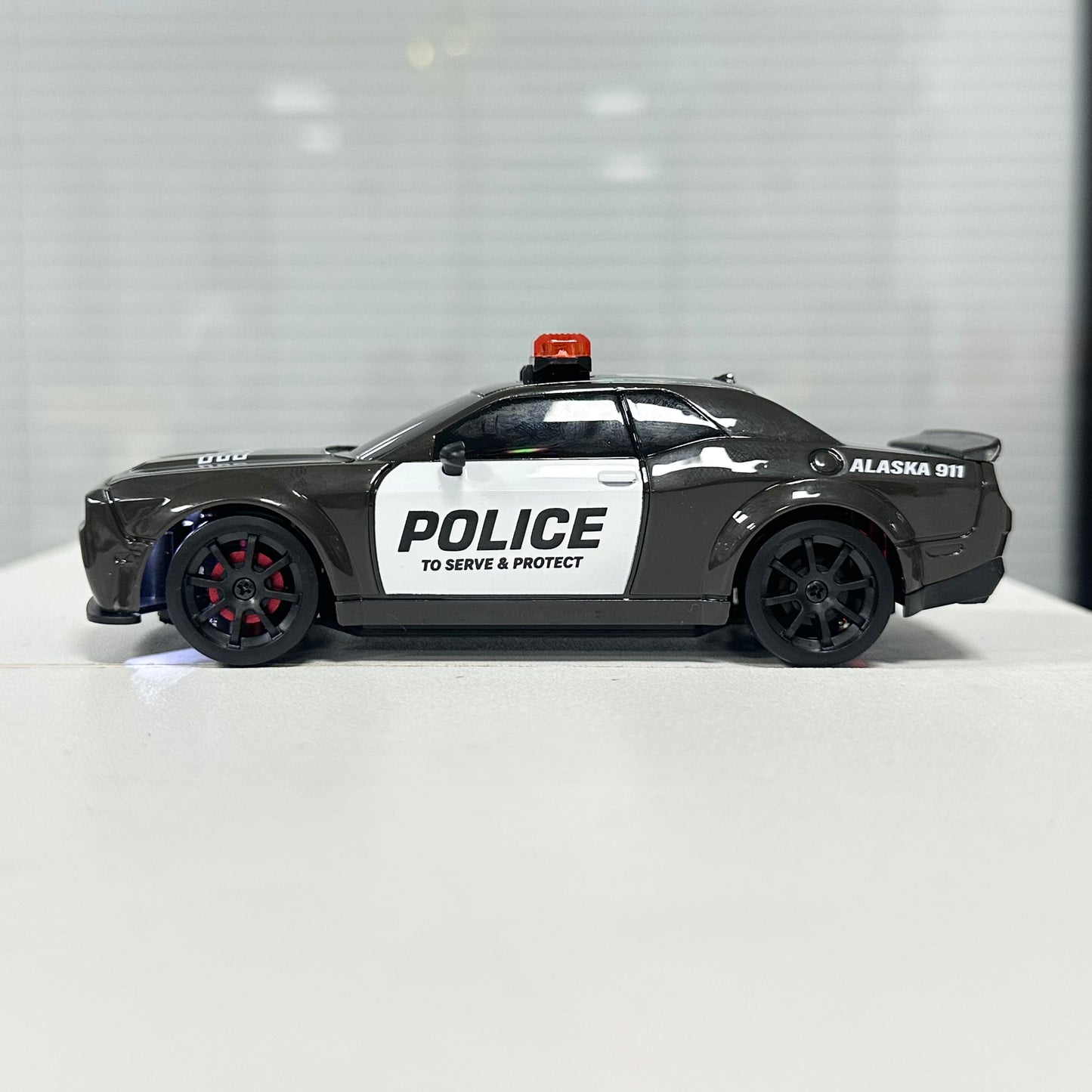 A toy model of a black and white police car with 'POLICE' and 'ALASKA 911' markings, displayed on a white surface against a blurred gray background.