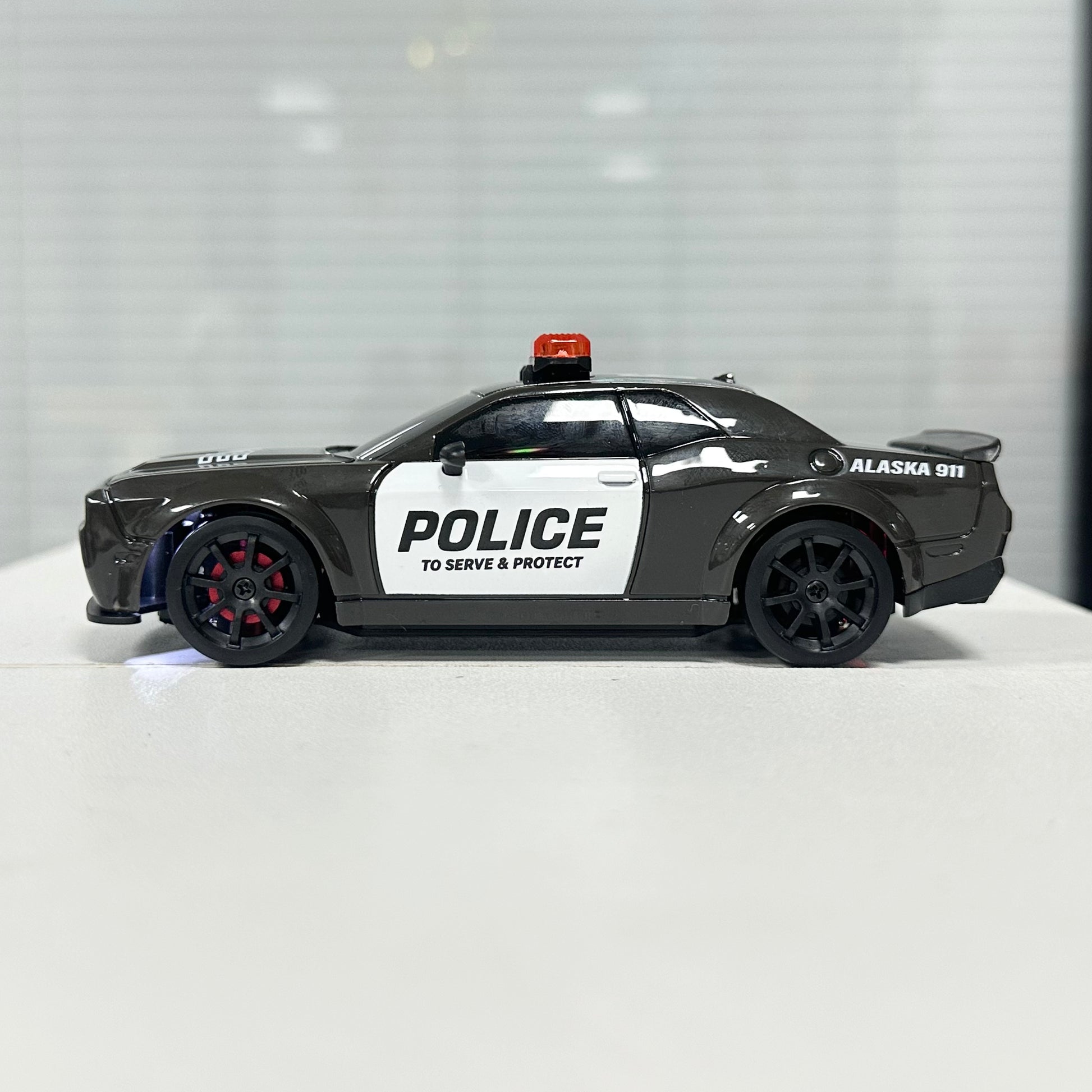 A toy model of a black and white police car with 'POLICE' and 'ALASKA 911' markings, displayed on a white surface against a blurred gray background.