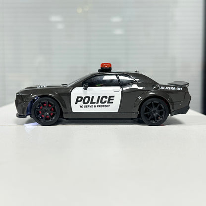 A toy model of a black and white police car with 'POLICE' and 'ALASKA 911' markings, displayed on a white surface against a blurred gray background.